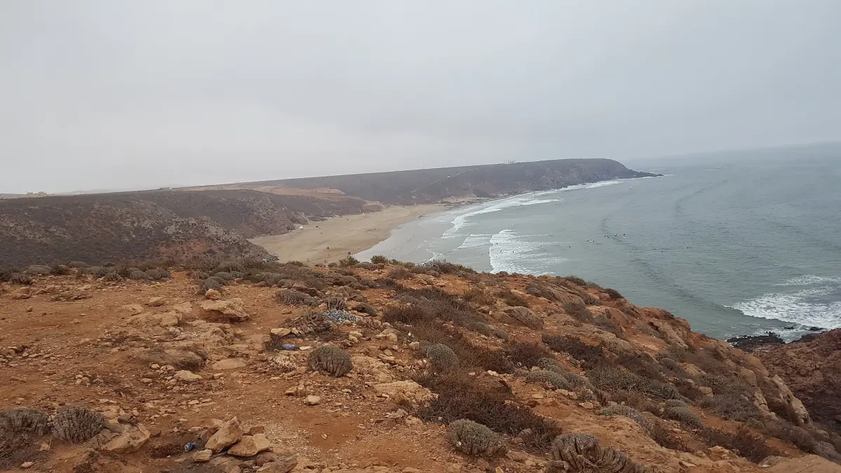 Wide view of Mirleft's coast with a sandy beach below brown cliffs and a hazy sea