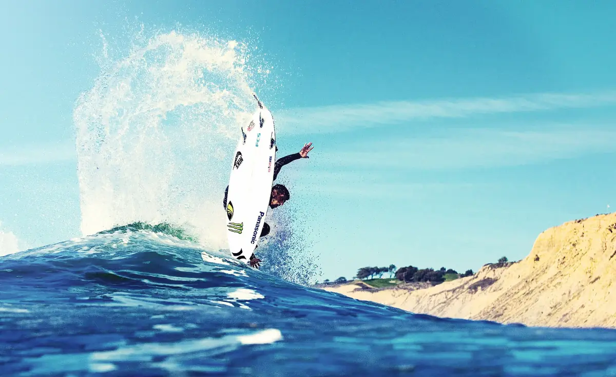 Surfer launching off a breaking wave with spray against blue water and a sandy cliff